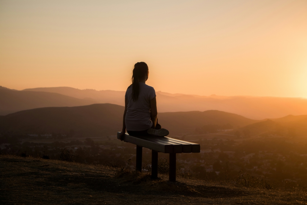 Uma pessoa sendata em um banco de madeira, olhando o horizonte e montanhas ao fundo com um por do sol alaranjado. 
Lifelong learning
